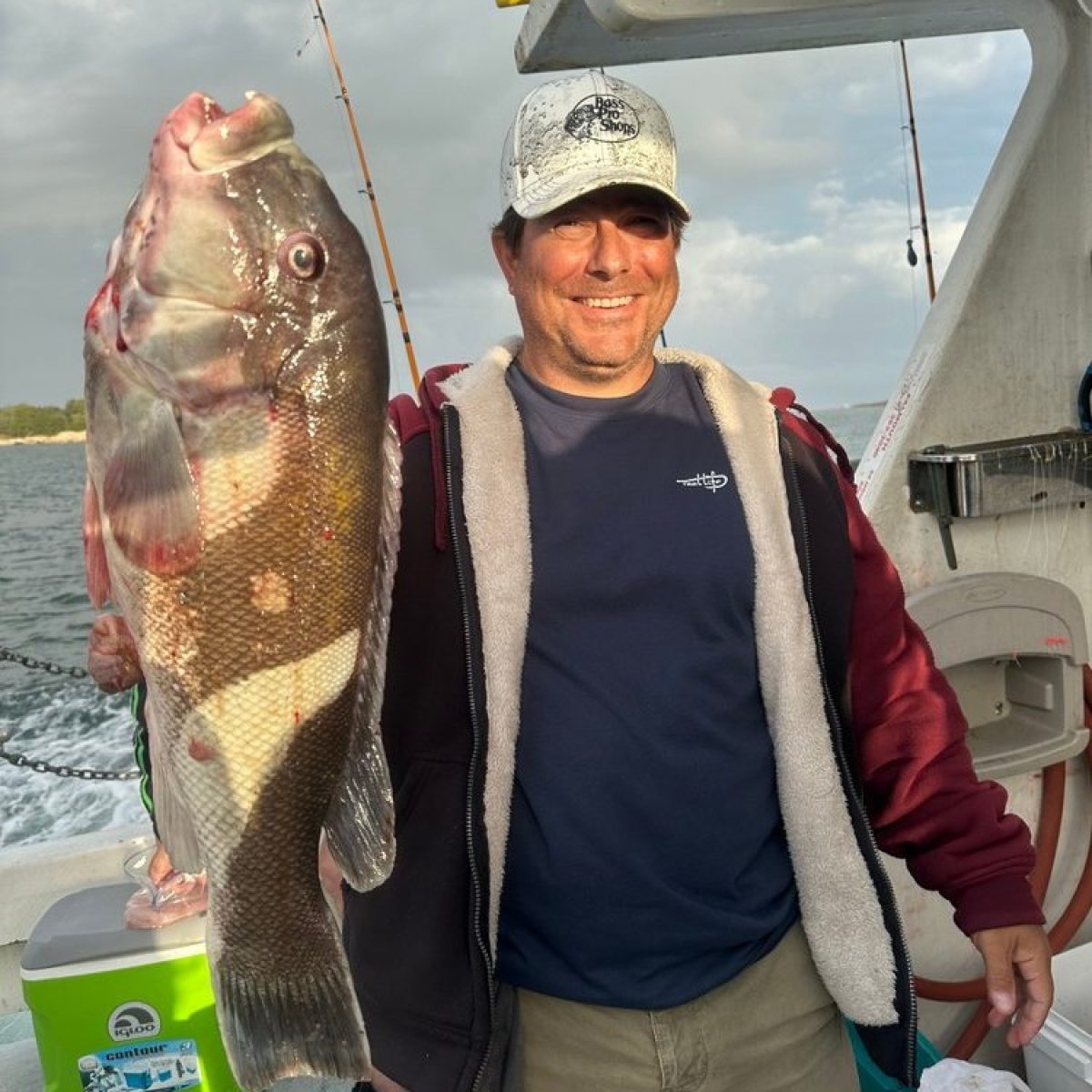 Man on a boat holding a large fish, smiling, with fishing rods in the background.