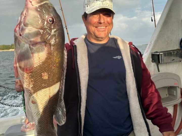 Man on a boat holding a large fish, smiling, with fishing rods in the background.