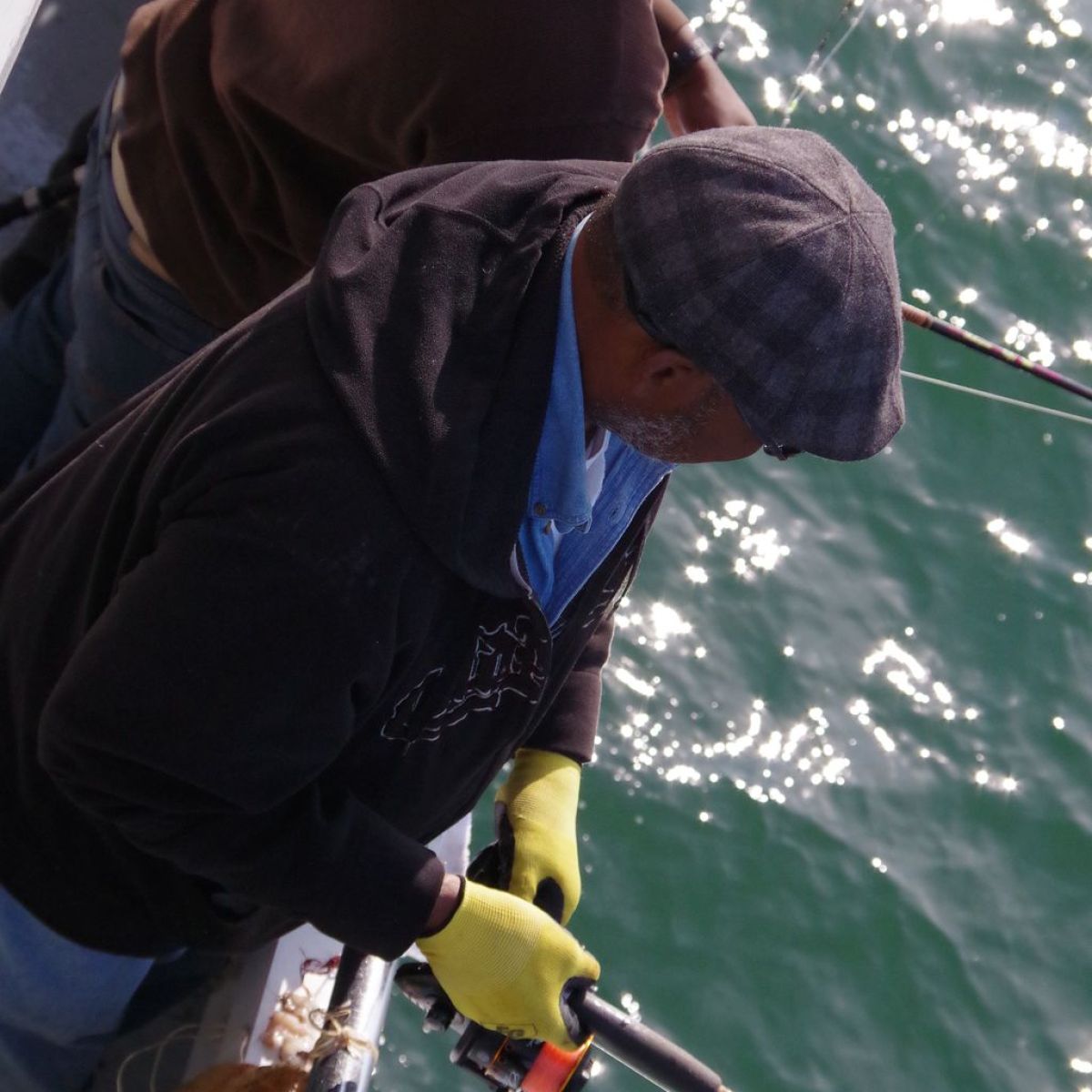 Two people fishing from a boat, one wearing a cap and yellow gloves, with water in the background.
