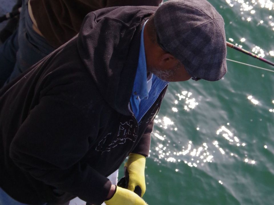 Two people fishing from a boat, one wearing a cap and yellow gloves, with water in the background.