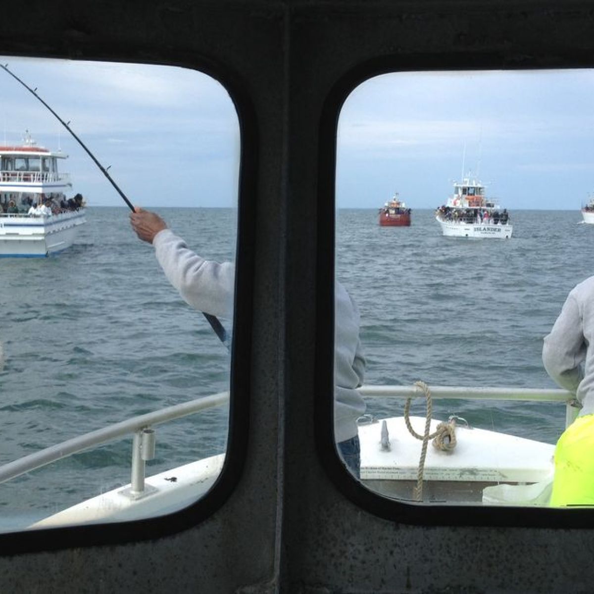 View through boat window of person fishing, catching a fish, and ships in the distance.