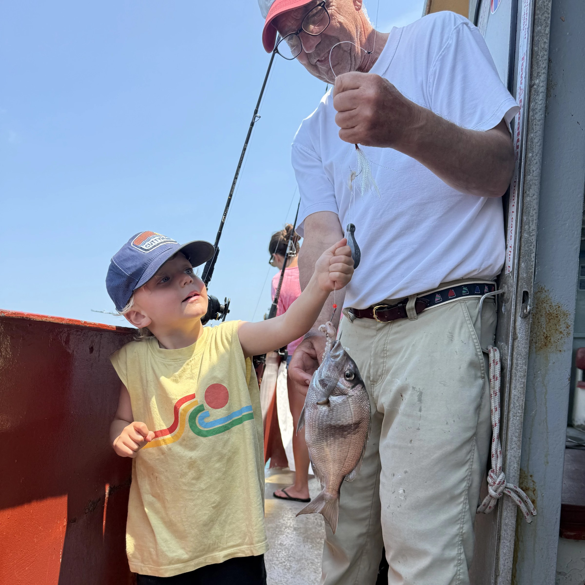 Child and adult hold a fish on a boat deck under a sunny sky.